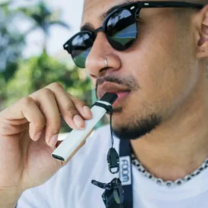 A young bearded man holding a pod system vape kit up to his mouth.